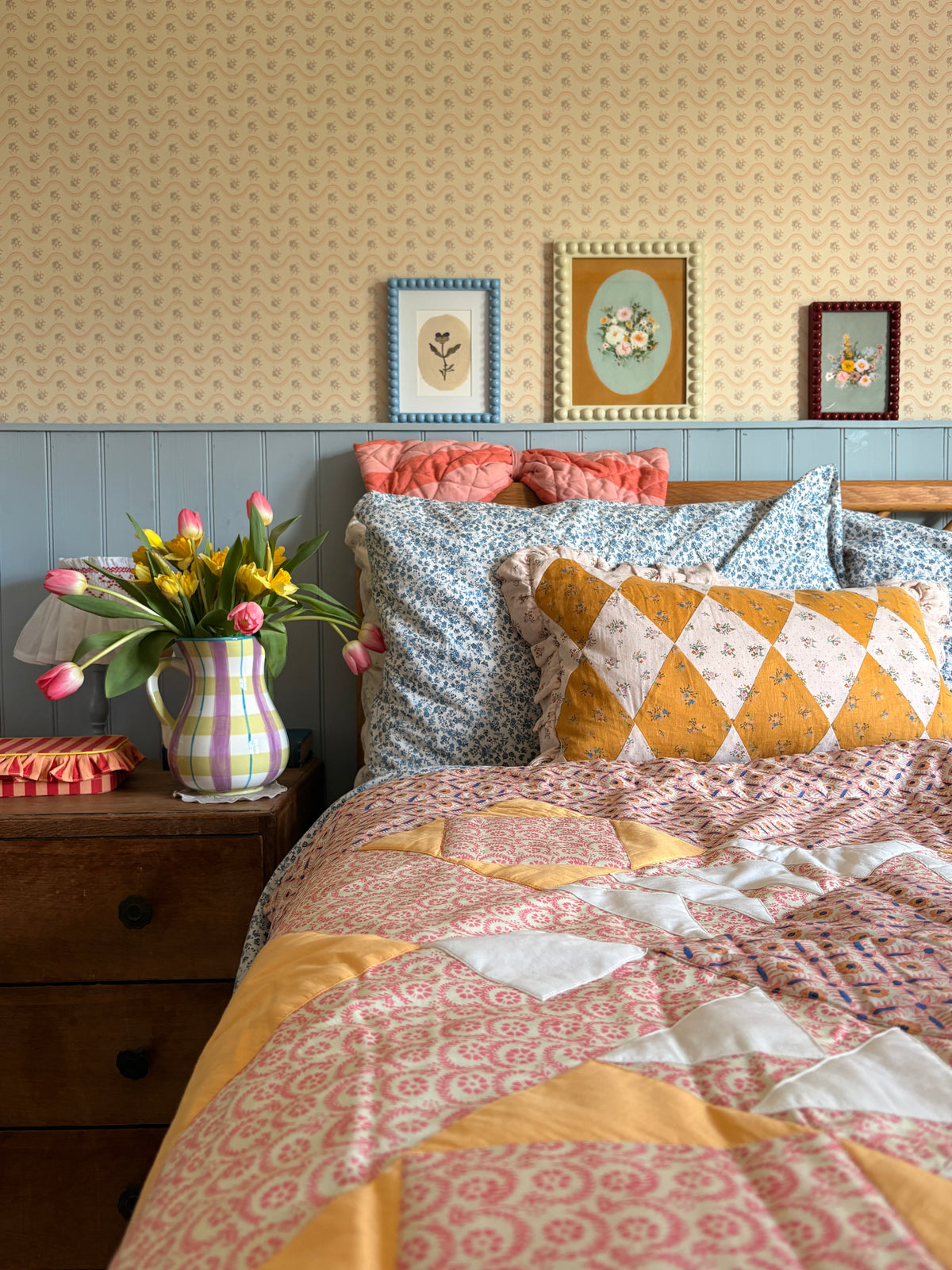 Bed with patterned bedding and pillows, floral arrangement, and framed pictures on a wall.